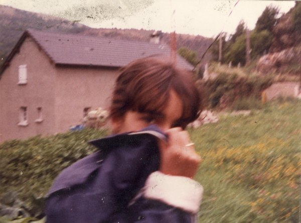 Femme cachant son visage avec un pan de blouson, en arrière-plan étendue d’herbe et maison.