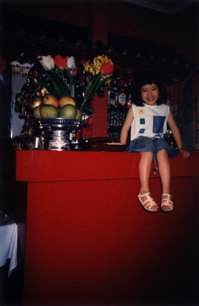 Petite fille riant assise de face sur un comptoir, vase de fleurs et corbeille de fruits en arrière-plan.