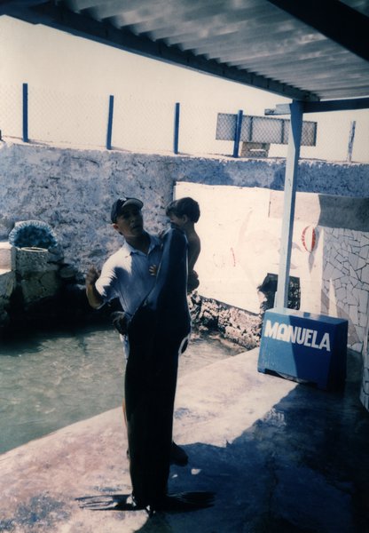 Homme portant un enfant près d'une piscine, enseigne « MANUELA » visible.ᴬᴵ