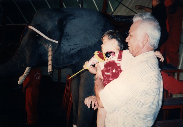 Homme âgé de profil avec un enfant dans les bras devant un éléphant.