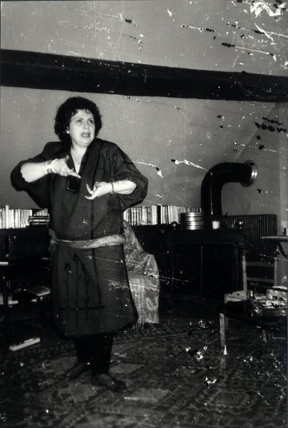 Femme parlant avec les mains dans un salon avec bibliothèque. Image très abîmée. Noir et blanc.ᴬᴵ