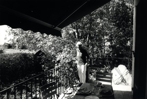 Homme debout sur une terrasse ombragée avec table dressée. Noir et blanc.ᴬᴵ