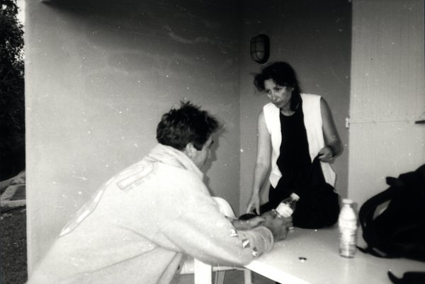 Homme assis à une table extérieure et femme debout à ses côtés. Noir et blanc.ᴬᴵ
