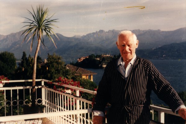 Homme âgé en robe de chambre rayée sur un balcon avec vue sur un lac et des montagnes.ᴬᴵ