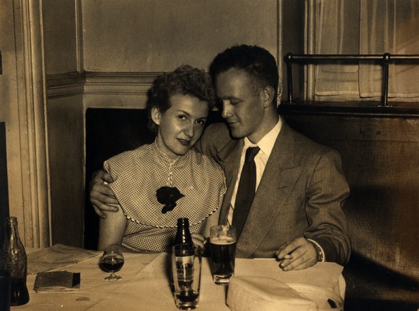 Couple attablé dans un restaurant, l'homme le bras autour de la femme. Noir et blanc.ᴬᴵ