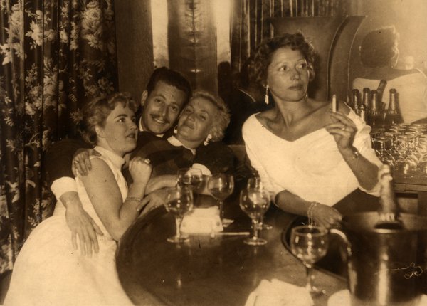 Quatre personnes à une table avec verres de vin, une femme fumant. Noir et blanc.ᴬᴵ