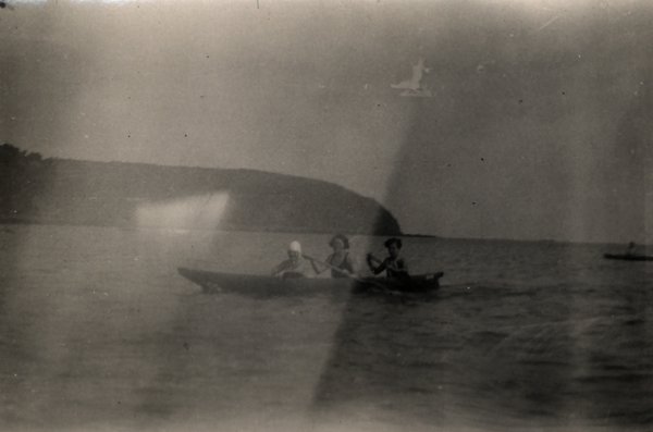 Trois personnes dans un canoë sur une mer calme, promontoire en arrière-plan. Noir et blanc.ᴬᴵ