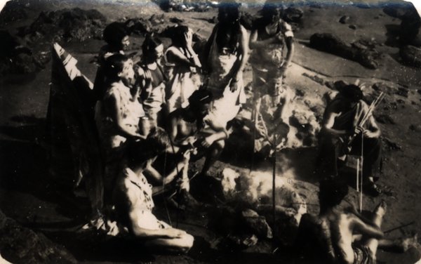 Groupe de personnes déguisées réunies autour d'un feu sur une plage rocheuse. Noir et blanc.ᴬᴵ
