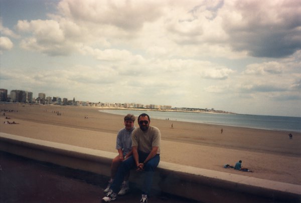 Un homme et un enfant assis sur un muret face à une plage.ᴬᴵ