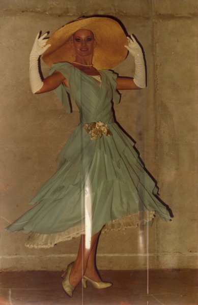 Grande femme avec capeline, robe à volants verts amande et longs gants blancs, prend la pose.