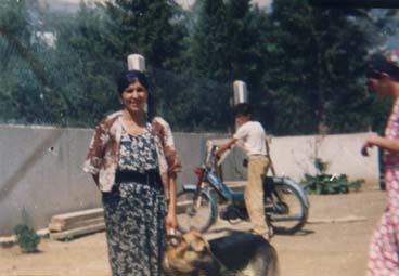 Femme brune en robe fleurie debout en extérieur avec un berger allemand, homme avec une mobylette en arrière-plan.