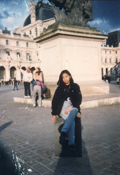 Jeune femme pose assise devant une statue sur l’esplanade de la Grande Pyramide du Louvre.