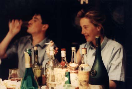 Homme brun et femme blonde avec une cigarette à la main côte à côte à une table où sont posées des bouteilles.