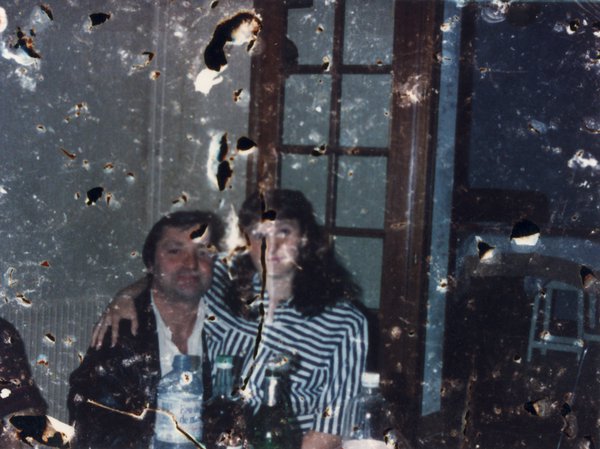 Femme brune à longs cheveux assise sur les genoux d’un homme brun attablé, bouteilles en avant-plan. Image très abîmée.