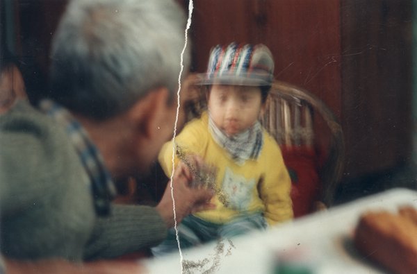 Petit enfant avec un sweat jaune et une casquette, homme en avant-plan. Image déchirée, reconstituée.