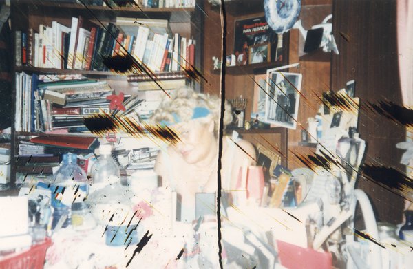 Femme blonde à bandeau turquoise assise derrière un bureau en désordre, étagères remplies de livres en arrière-plan.