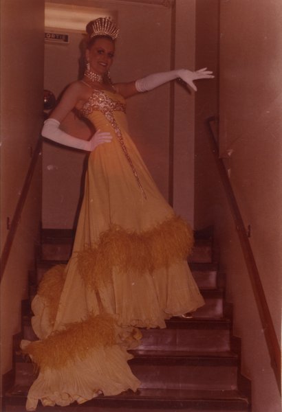 Femme en robe longue à frou frou, gants blancs et diadème, pose en haut d’un escalier.