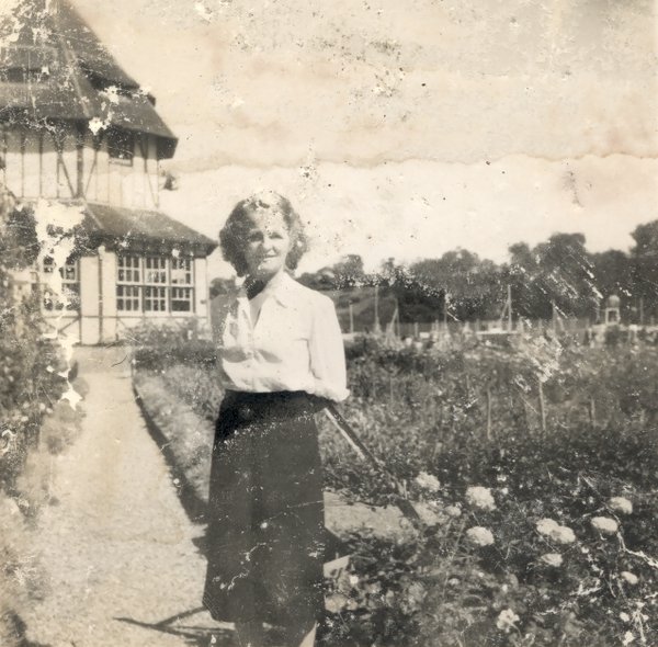 Femme blonde en jupe sombre debout dans un jardin, massifs de fleurs en arrière-plan.