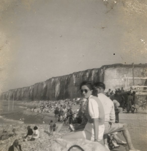 Femme en jersey blanc assise sur un parapet au bord de la mer, falaises en arrière-plan.