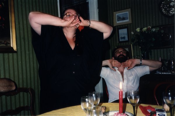 Femme criant ou chantant les mains devant la bouche, un homme derrière elle à table.ᴬᴵ