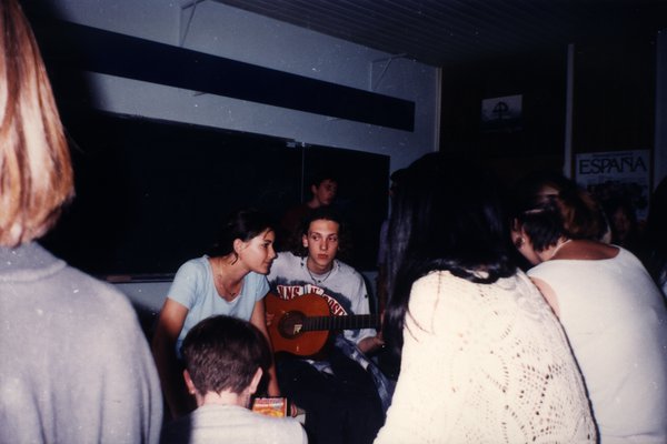 Adolescents assis autour d'un joueur de guitare, affiche « España » au mur.ᴬᴵ