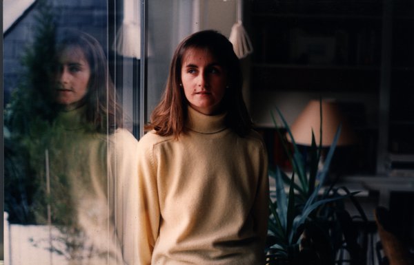 Jeune femme debout près d'une fenêtre avec son reflet visible, plantes vertes à proximité.ᴬᴵ