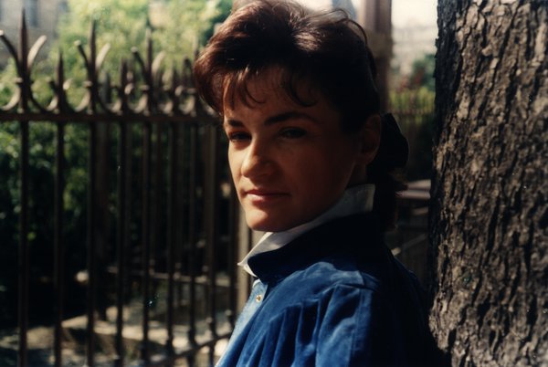 Portrait d'une femme en veste bleue appuyée contre un arbre, regardant l'objectif.ᴬᴵ