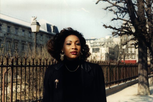 Femme aux cheveux bouclés debout près d'une grille en fer forgé dans une rue.ᴬᴵ