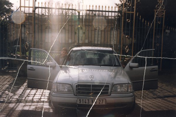 Mercedes grise métallisée toutes portes ouvertes devant une grille en fer forgé et un enfant, belle maison en arrière – plan.