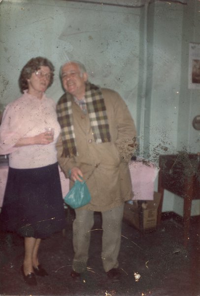 Femme un verre à la main avec un homme âgé avec un sac plastique vert à la main, une écharpe à carreaux autour du cou.