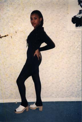 Jeune fille pose en pull et collant noir de profil avec des chaussures à talon devant un mur. `