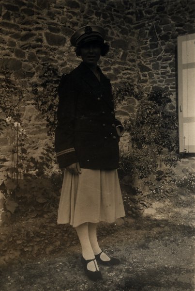 Femme en veste sombre et casquette devant un mur en pierre. Noir et blanc.ᴬᴵ