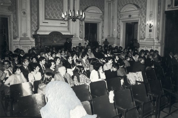 Public assis sur une chaise dans une salle à lambris et lustres. Noir et blanc.
