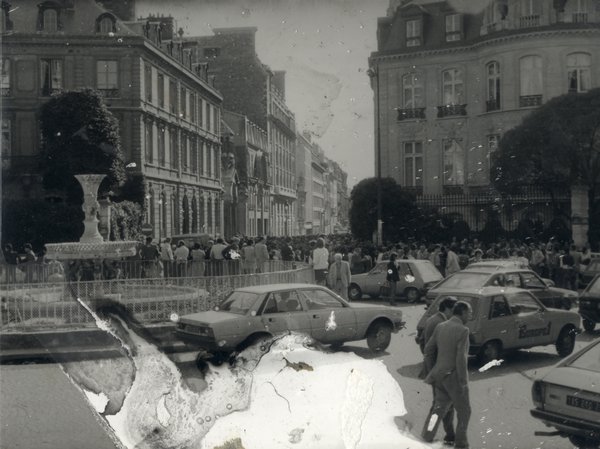Place avec fontaine, passants et voitures.