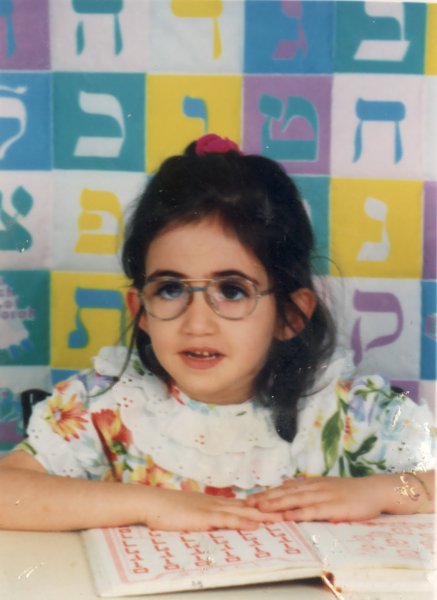 Petite fille brune à lunette devant une table, lettres hébraïques  de couleur en arrière-plan.