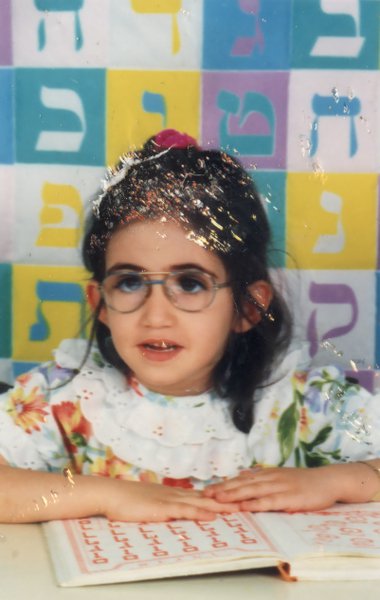 Petite fille brune à lunette devant une table, lettres hébraïques  de couleur en arrière-plan.