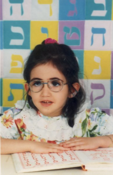 Petite fille brune à lunette devant une table, lettres hébraïques  de couleur en arrière-plan.