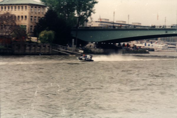 Bateau de course passant sous un pont, immeubles en arrière-plan.ᴬᴵ