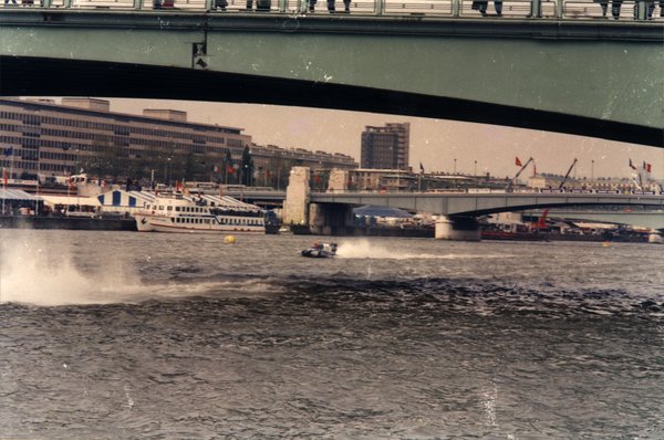 Bateau de course sur une rivière en ville, sous un pont, avec bateaux de tourisme amarrés.ᴬᴵ