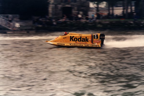 Bateau de course Kodak sur une rivière devant un bâtiment en pierre.ᴬᴵ