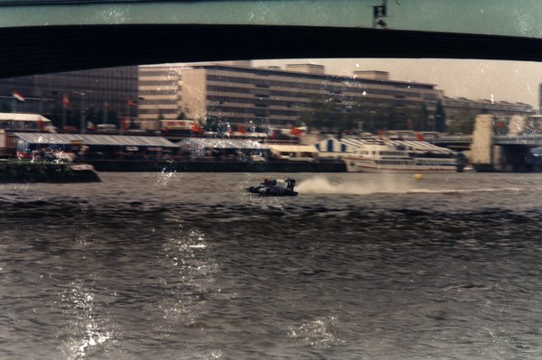 Bateau de course sous un pont, avec bateaux de tourisme et immeubles en arrière-plan.ᴬᴵ