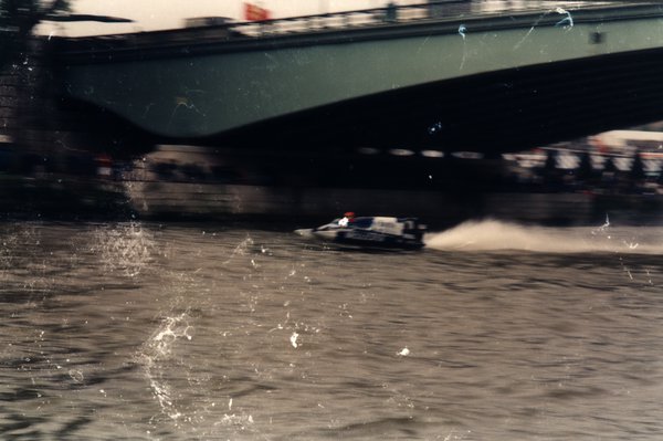 Bateau de course sous un pont, sillage d'écume et foule sur la rive.ᴬᴵ