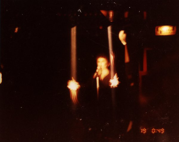 Transformiste en Edith Piaf accompagné d’un homme déguisé en arlequin sur une scène face à un public.