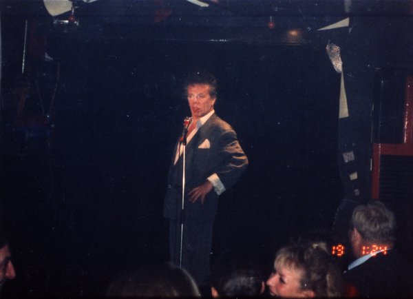 Chanteur en costume gris avec un micro sur une scène face à un public.