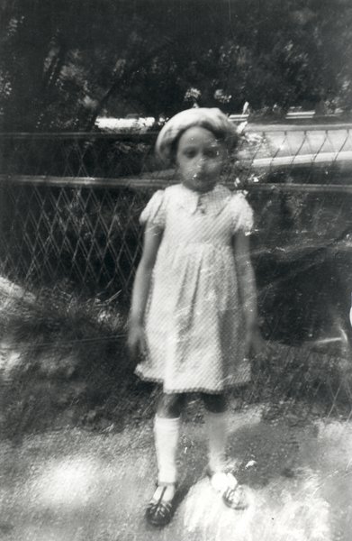 Petite fille en robe et chapeau debout devant un grillage. Noir et blanc.