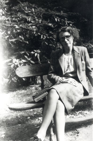 Femme en jupe et veste à lunettes assise sur un banc. Noir et blanc.