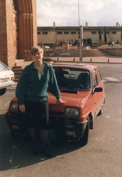 Femme rousse à cheveux courts debout devant une renaud 5 rouge sur une place avec une église.